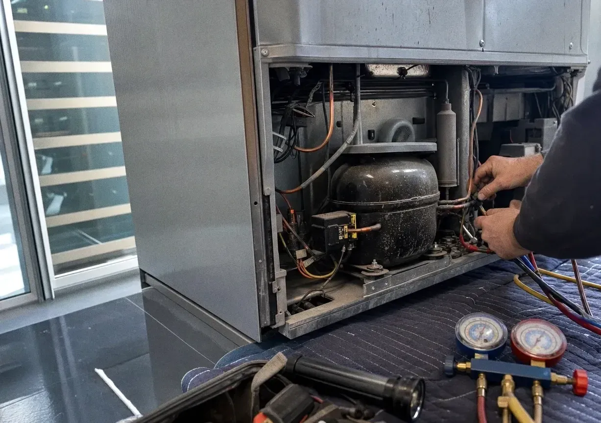 Rear access panel removed from a glass-doored cabinet, exposing the sealed system with gauges on the floor beside it